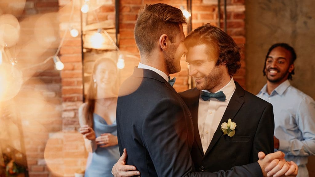 Two grooms share a romantic first dance at their wedding, surrounded by smiling guests and warm lighting.