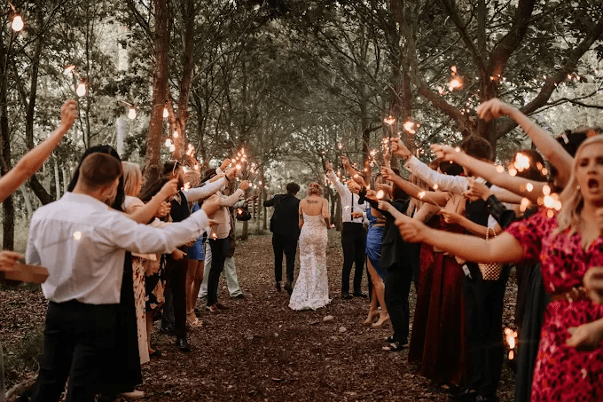 ride and groom walking through woodland as wedding guests hold sparklers in celebration.