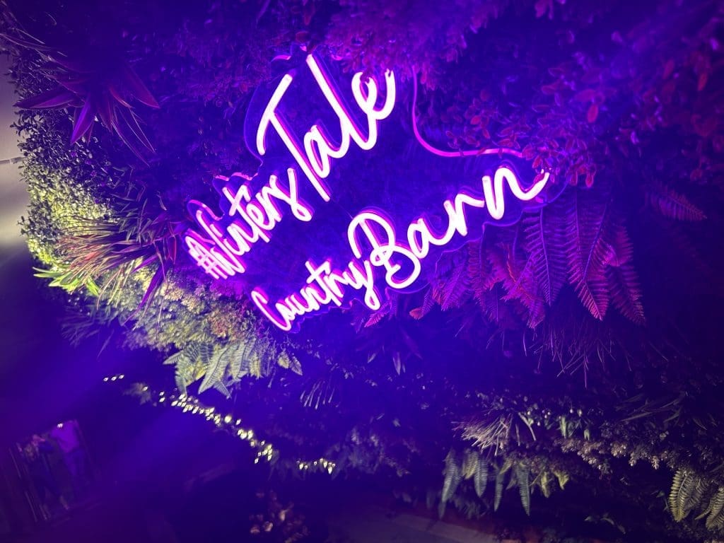 Purple neon Winter’s Tale Country Barn sign surrounded by greenery.